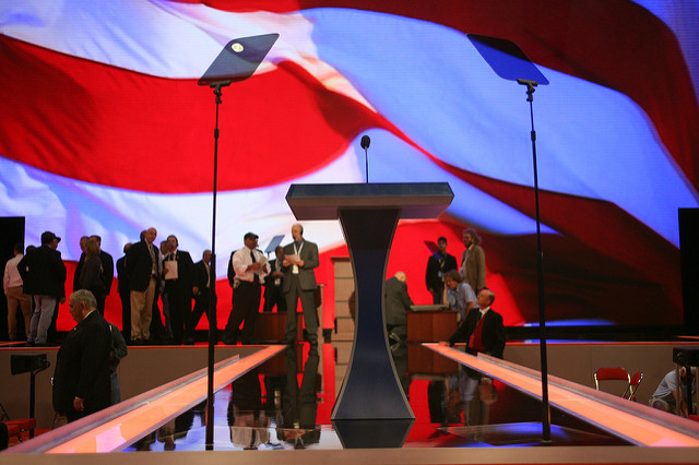 Low-angle view of a political stage podium set up against a large, blurred backdrop of the American flag.